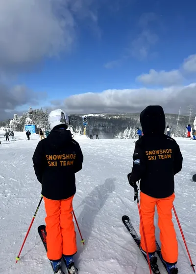 Two Snowshoe Ski Team racers looking out over snowy trees and blue sky from the top of the slope