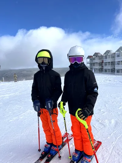 Two young skiers in goggles and orange race pants posing on a groomed slope under a blue sky