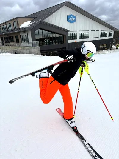 A young skier in orange pants balancing on one ski with the other held up, in front of the Shavers Center