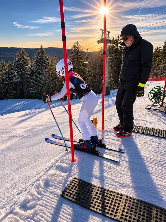 A young Snowshoe ski team racer in the start gate beside a coach at sunset