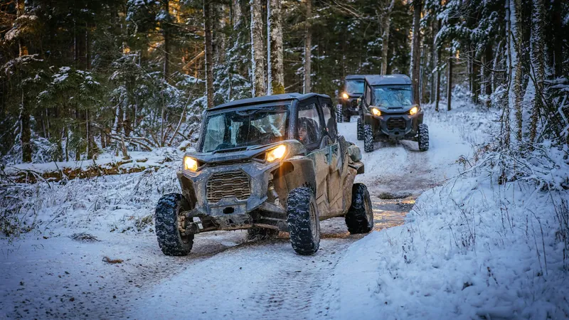 Two side-by-side off-road vehicles with headlights on, driving a snowy trail through the forest
