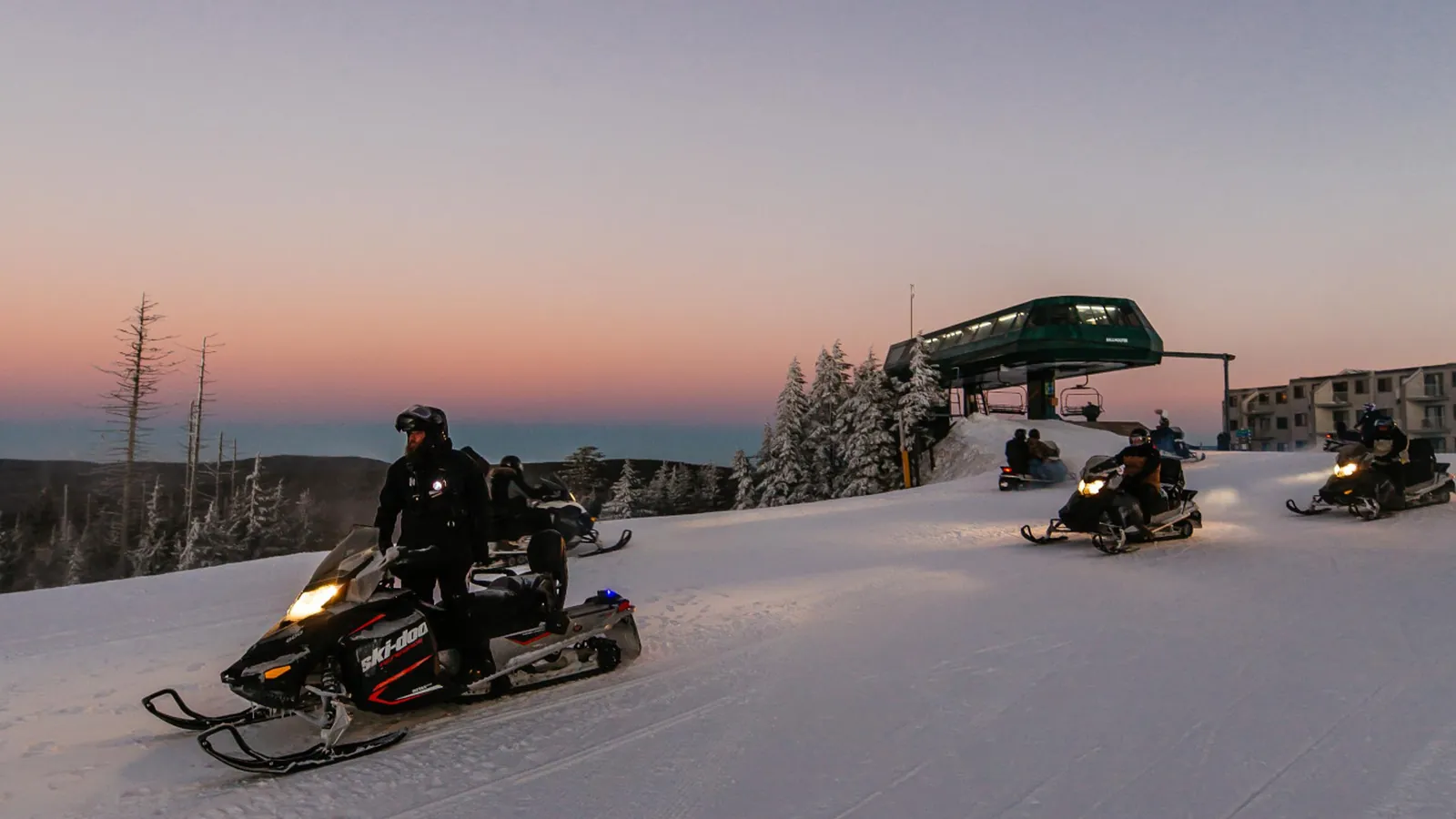Riders on snowmobiles crossing a groomed slope at sunset, with a chairlift station behind them at Snowshoe