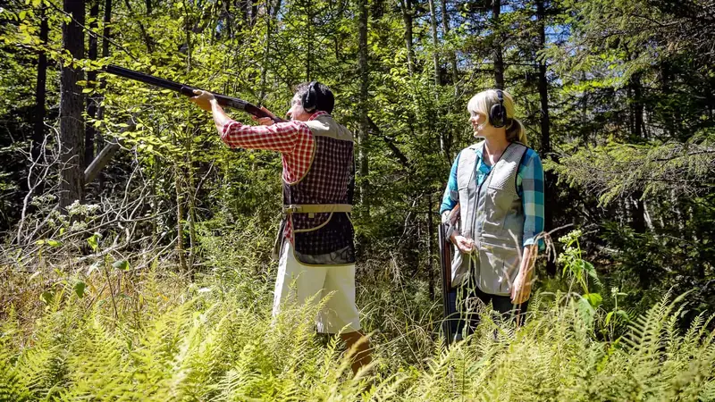 A shooter taking aim on the sporting clays course in a green forest at Snowshoe, with an instructor beside him