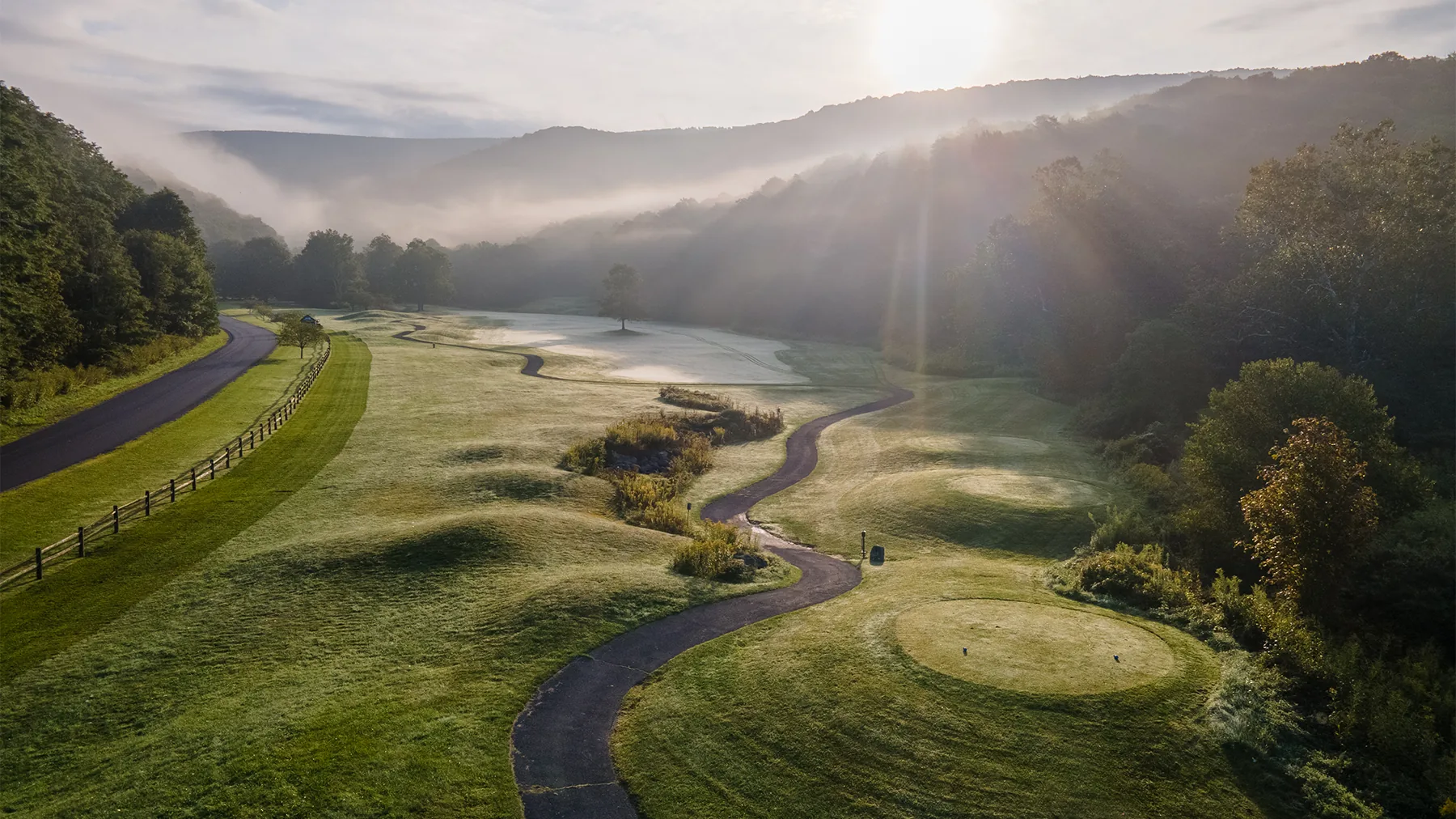 Misty sunrise over the Raven Golf Club, a valley course with a winding cart path, bunkers, and a putting green