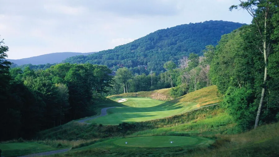 A Raven tee box looking up a fairway toward a forested mountain, with a sand bunker and a green in the foreground